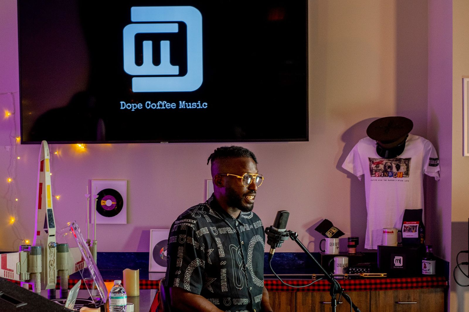 Creative Mike at the mic in the Dope Coffee Music studio, with the SPINACH tee on the wall